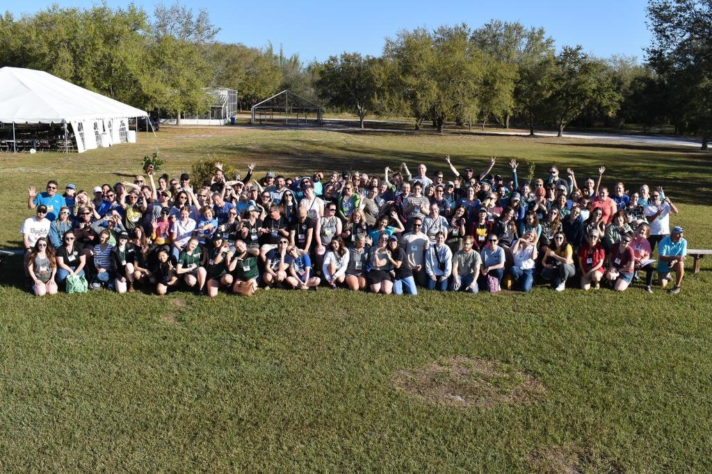 A large group of people sit in rows to pose for a picture. They are in a field with a white tent in the background